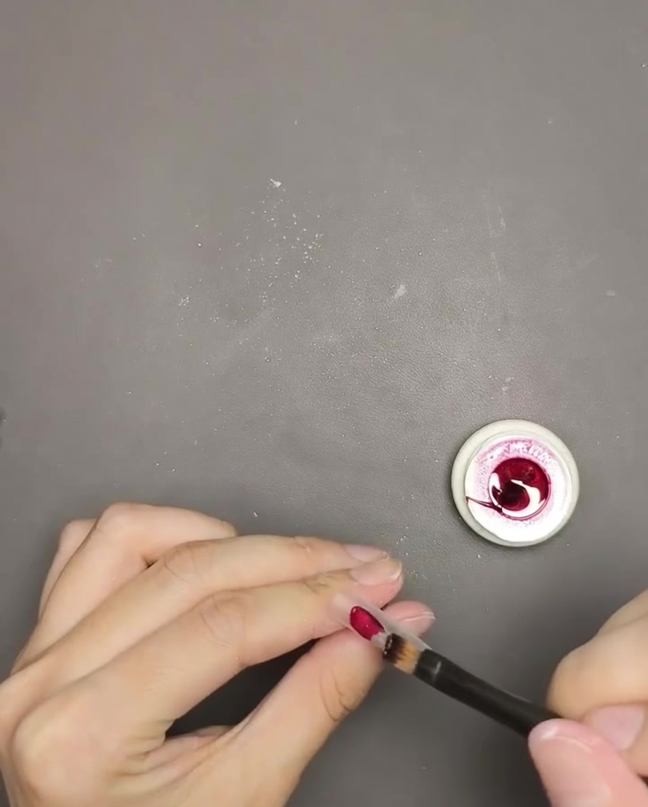 Primer plano de un pincel aplicando esmalte de gel ojo de gato rojo intenso sobre una punta de uña.