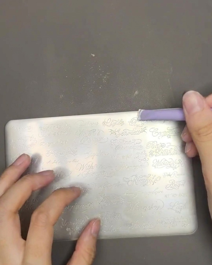 Primer plano que muestra esmalte de estampación blanco siendo extendido a través de una placa de metal con un raspador morado.