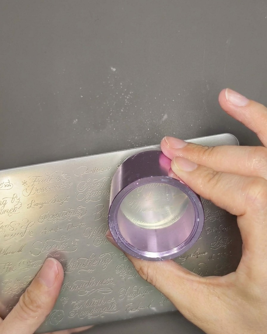 Vista de primer plano de un cabezal de estampador de gelatina transparente siendo sostenido en la mano, con una capa delgada de esmalte de estampación blanco aplicado sobre el diseño previamente recogido.