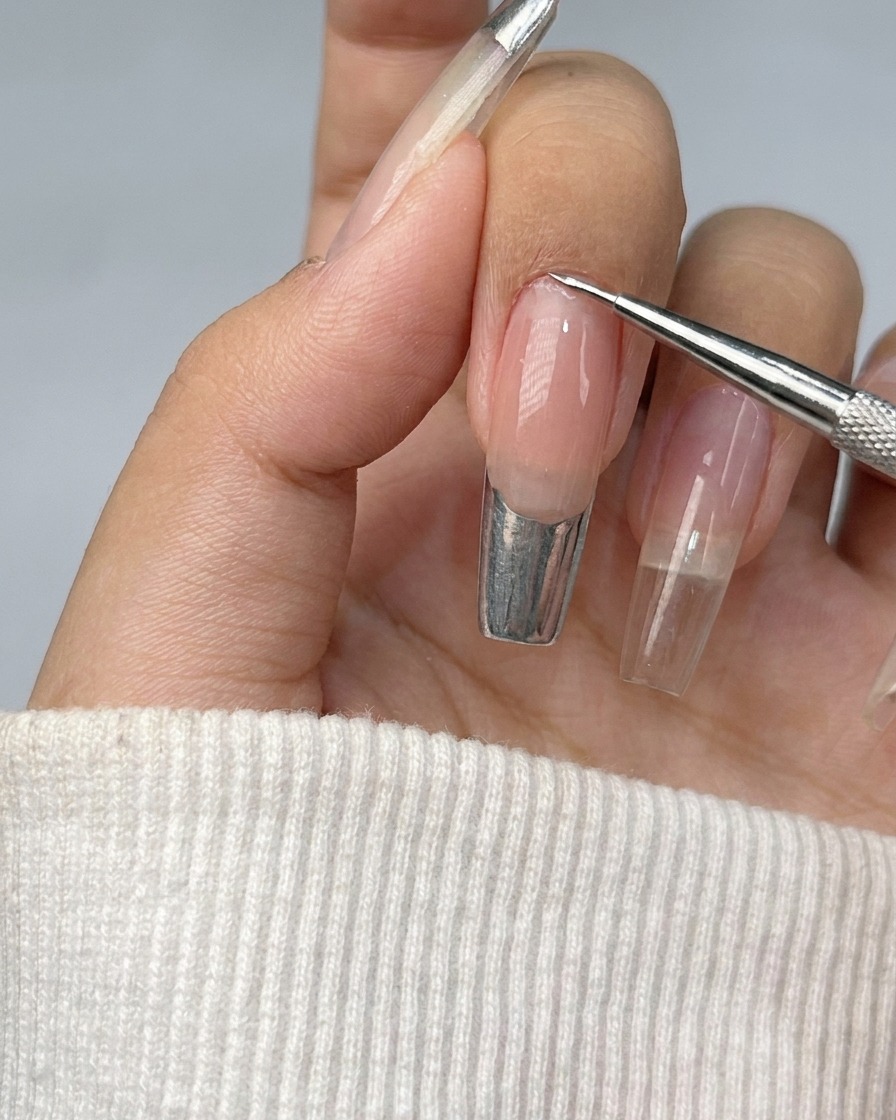 Una uña con una punta francesa siendo rellenada con gel metálico plateado brillante.