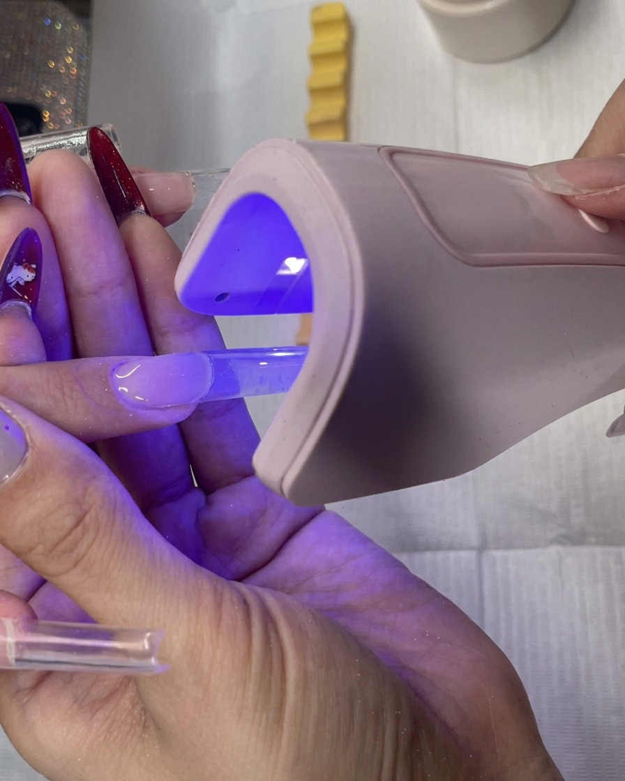 Un artista de uñas sosteniendo una pequeña lámpara LED rosa sobre una extensión de uña cuadrada transparente.