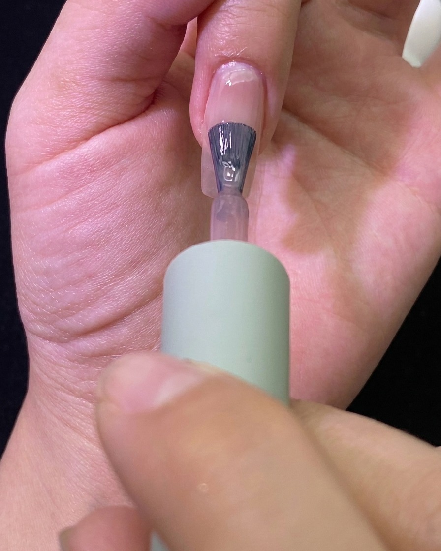 Primer plano de un pincel aplicando gel color nude transparente sobre una uña natural.