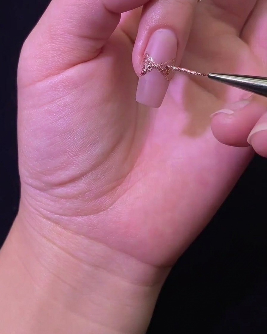 Primer plano de un pincel liner rellenando la forma del lazo con glitter sobre la superficie de la uña.