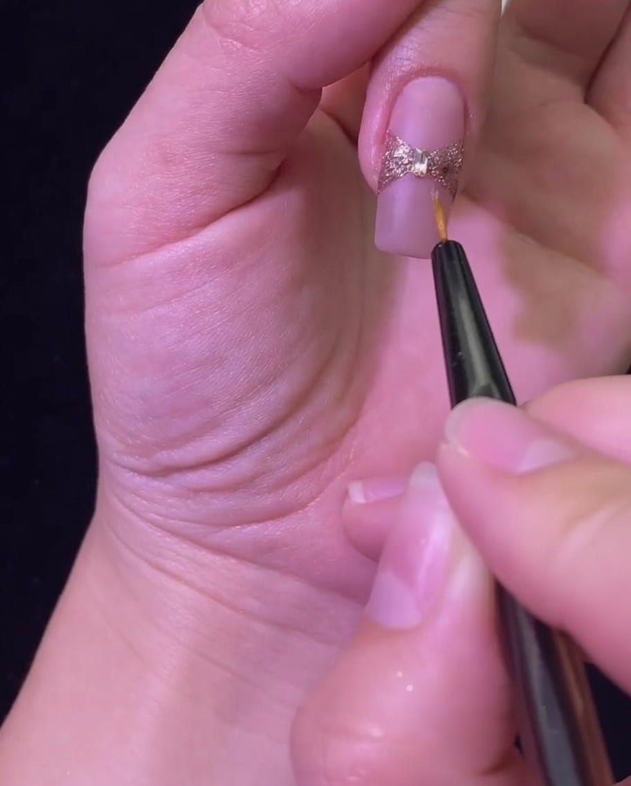 Un pincel liner aplicando top coat brillante sobre el lazo de glitter y el cristal central.