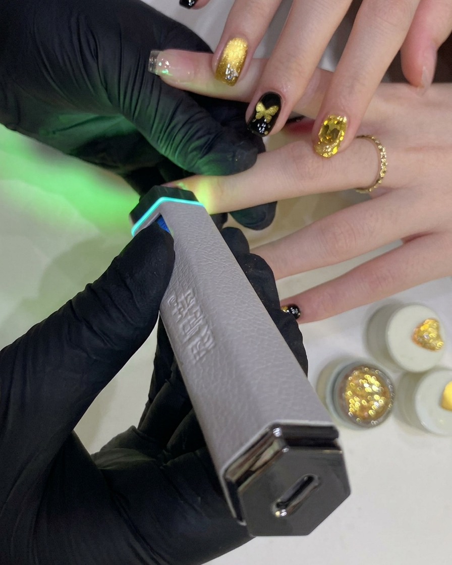 Una mano enguantada sostiene una lámpara UV/LED portátil sobre una uña pintada con esmalte de gel negro.