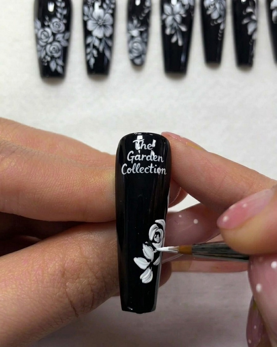 Vista de cerca de un artista de uñas añadiendo líneas blancas finas para representar tallos que se extienden desde el follaje en una uña pintada de negro.