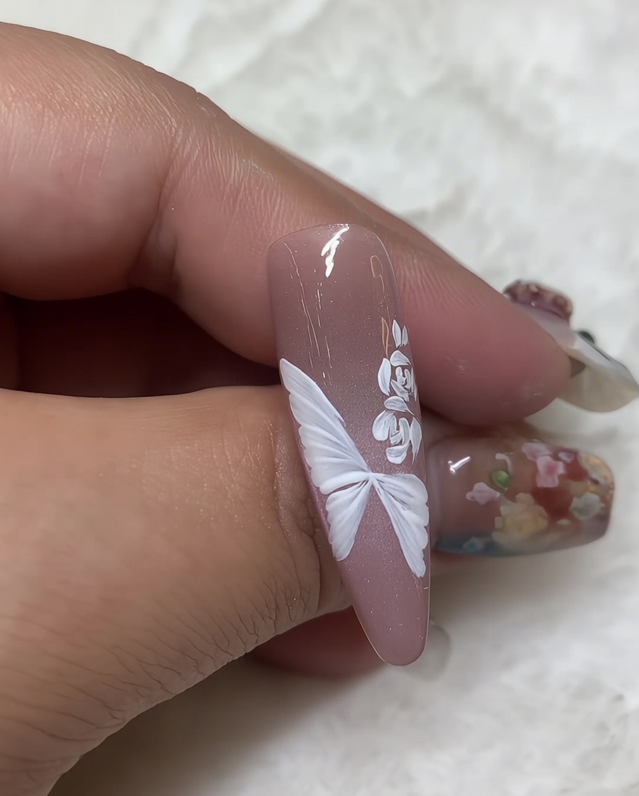 Primer plano de una uña con una rosa pintada en gel blanco y un pincel liner fino agregando hojas blancas pequeñas y detalladas a un lado del diseño floral.