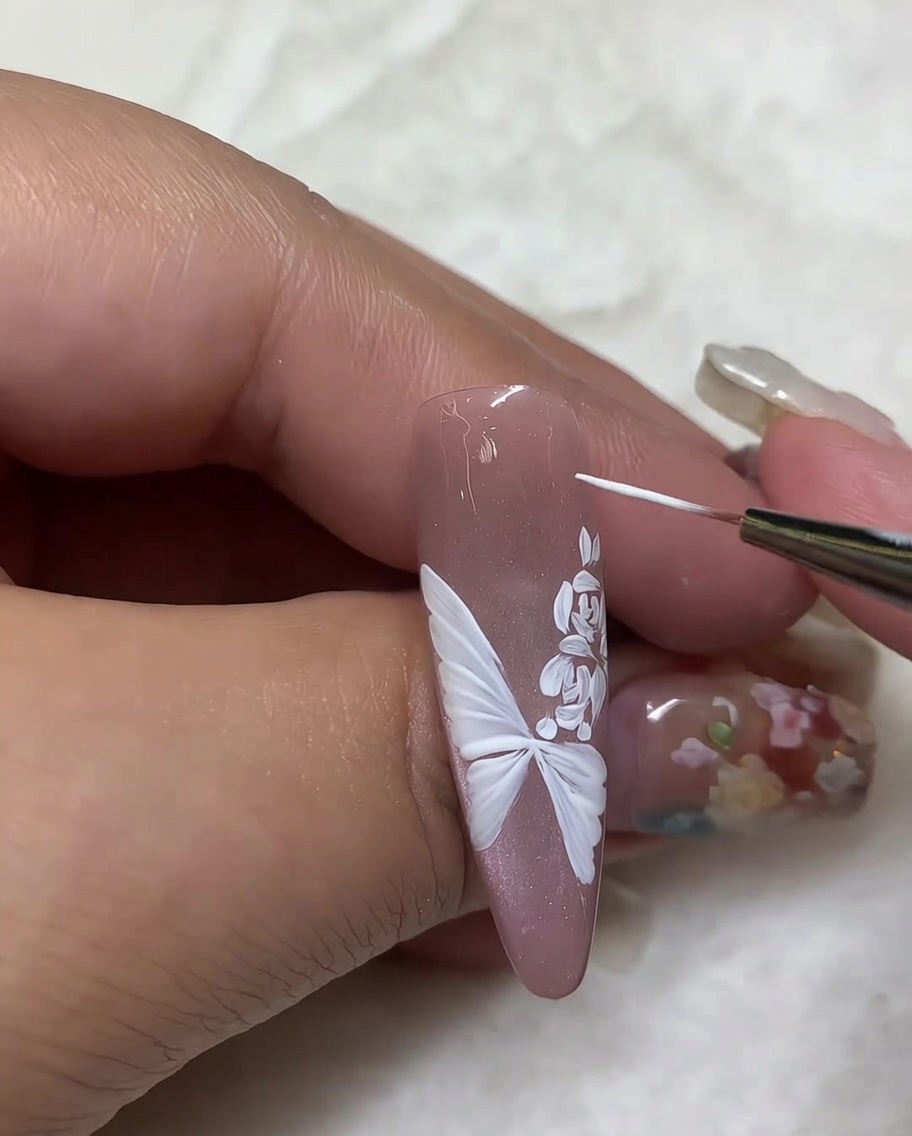 Vista detallada que muestra un pincel liner fino agregando pequeños puntos de luz blancos alrededor de un diseño de mariposa y rosa en una uña de tono nude.