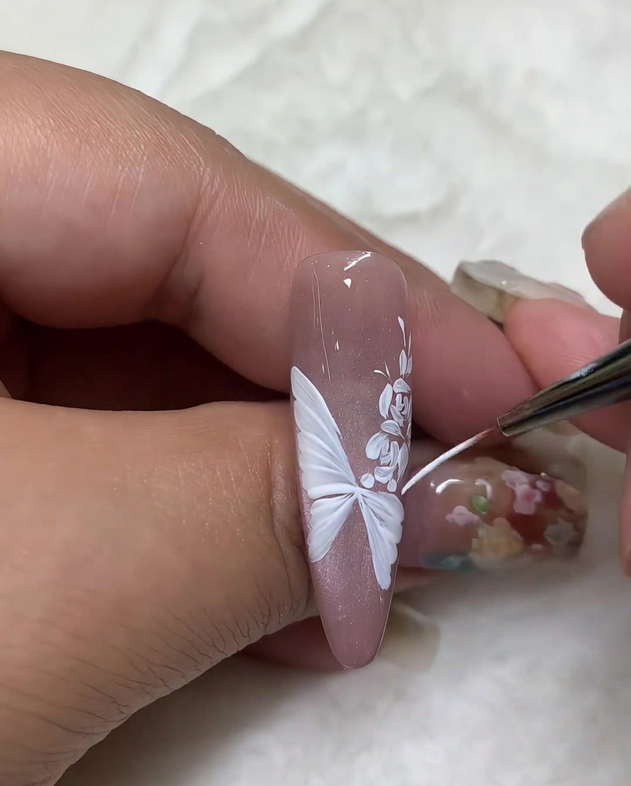 Primer plano de una uña mostrando un pincel liner fino trazando los bordes exteriores de un diseño de ala de mariposa blanca para refinar los detalles.