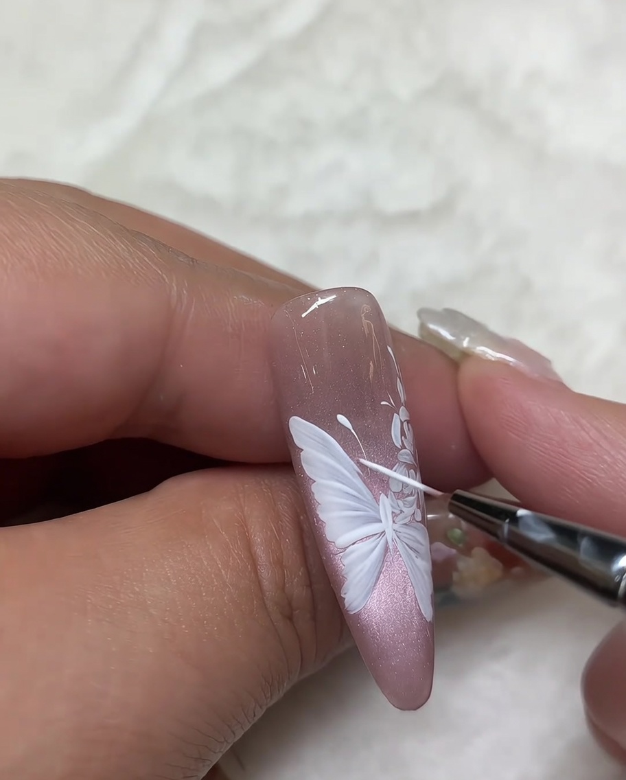 Toma cercana de una uña con un diseño de rosa blanca, usando un pincel liner fino para realizar los retoques finales en los detalles de los pétalos.