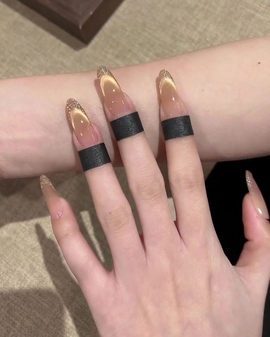 Primer plano de una mano usando un pincel delineador fino para pintar un borde de purpurina dorada en una uña cat-eye estilo francesa.
