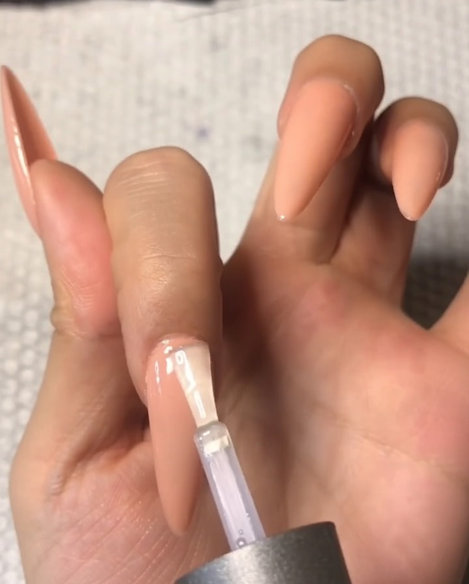 Primer plano de un pincel aplicando cuidadosamente un top coat templado transparente cerca del área de la cutícula de una uña del dedo medio con forma de almendra.