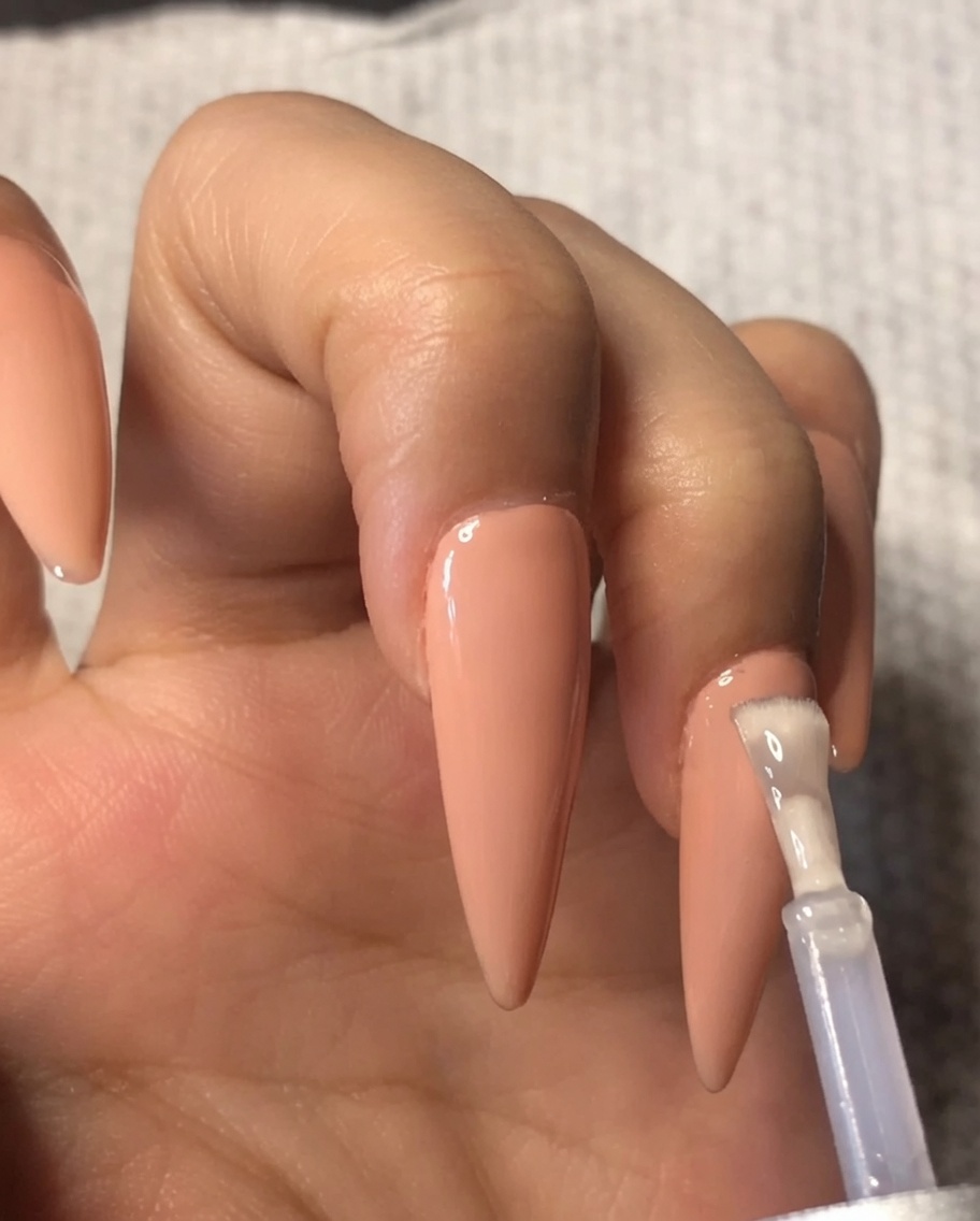 Un pincel aplicando una capa de top coat de gel transparente sobre un esmalte de gel nude en el dedo anular.