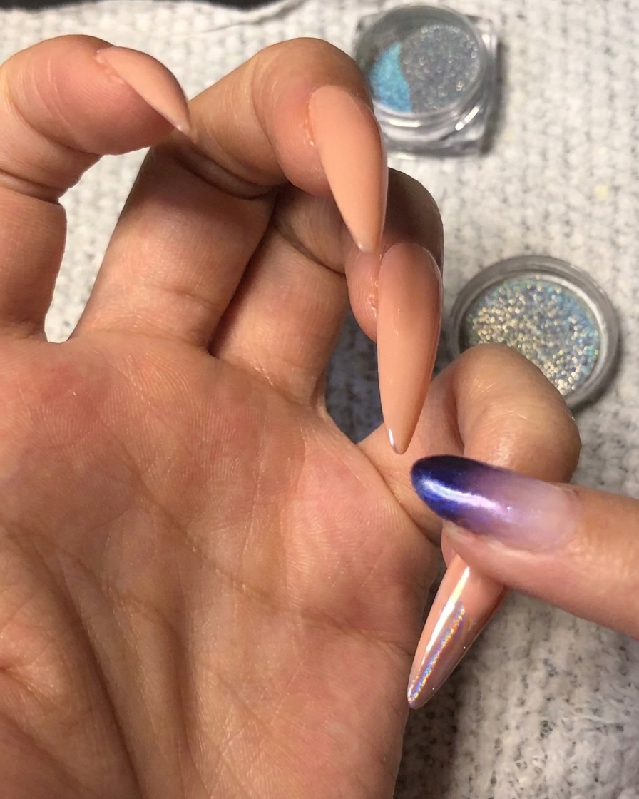 Un dedo presionando polvo cromado holográfico azul-púrpura vibrante sobre la punta de una uña con forma de almendra nude.
