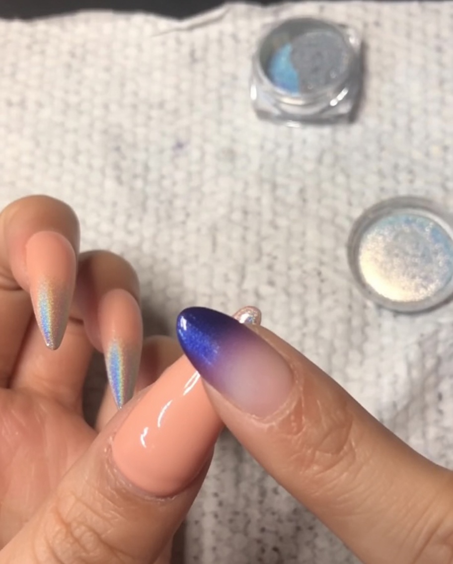 Primer plano de un dedo frotando polvo cromado holográfico sobre la punta de una uña meñique, creando un degradado ombré.
