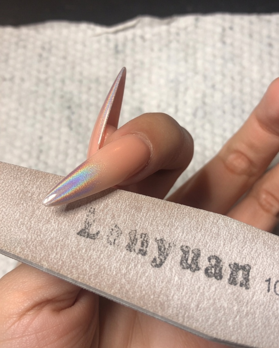 Una mano usando una lima de esponja fina para pulir suavemente el borde libre de una uña stiletto decorada con polvo cromado holográfico.