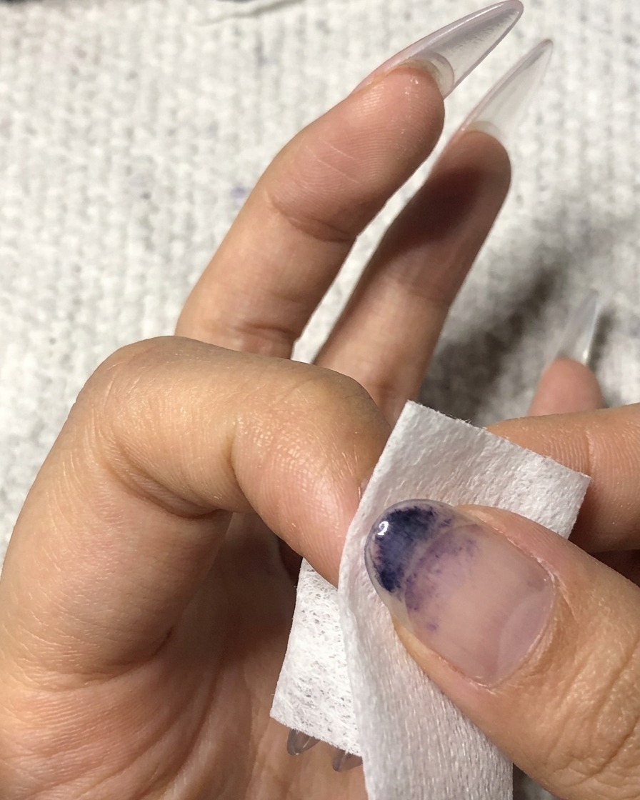 Una toallita sin pelusa siendo usada para limpiar la superficie de una uña del dedo anular manicurada.