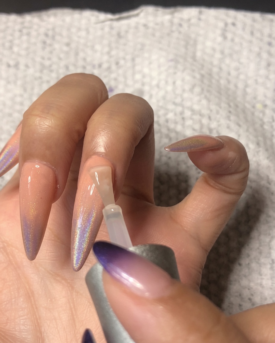 Un pincel de uñas aplicando un top coat de alto brillo al dedo anular, sellando el arte de uñas ombré holográfico.