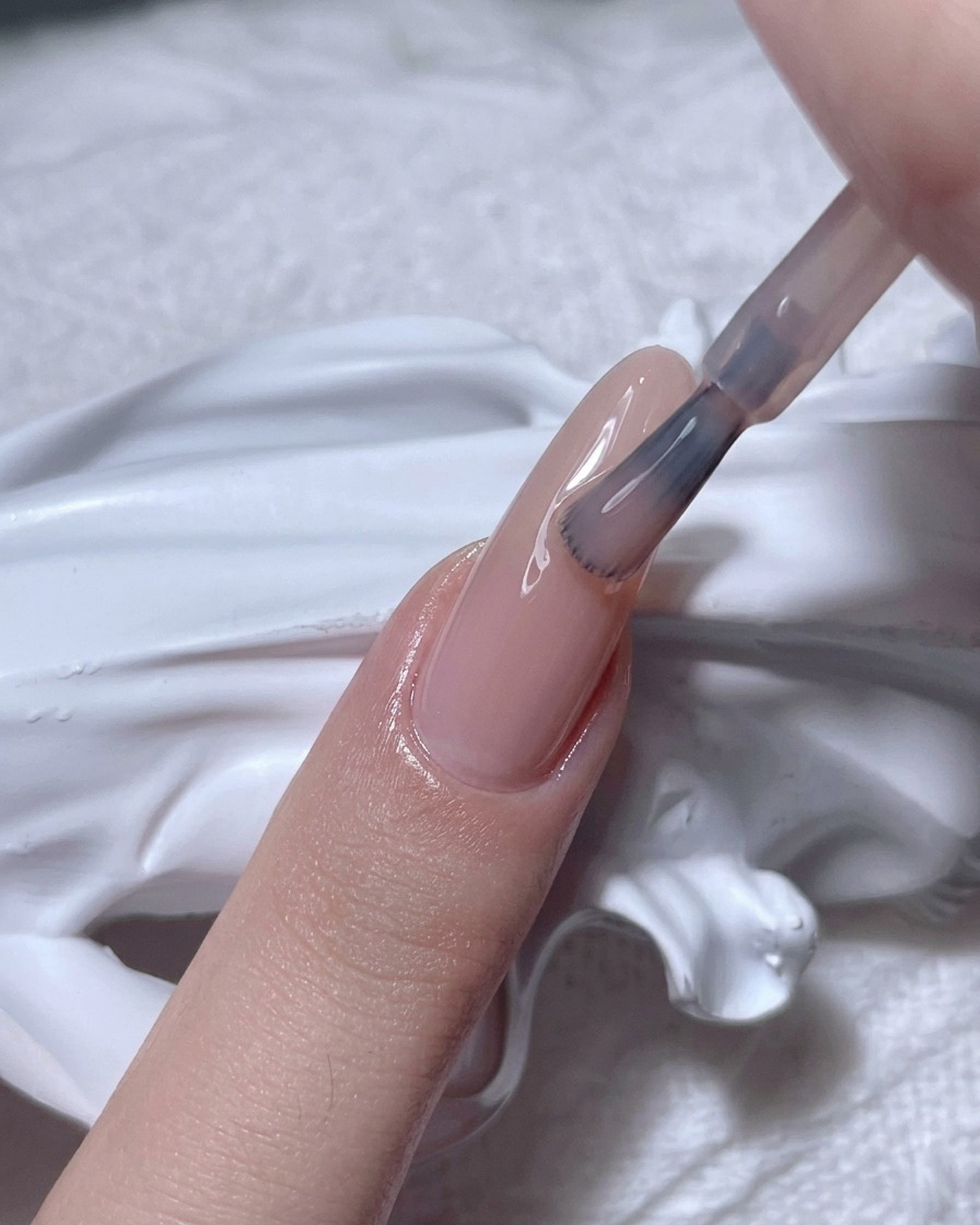 Un pincel aplicando esmalte de gel nude translúcido sobre una uña natural.