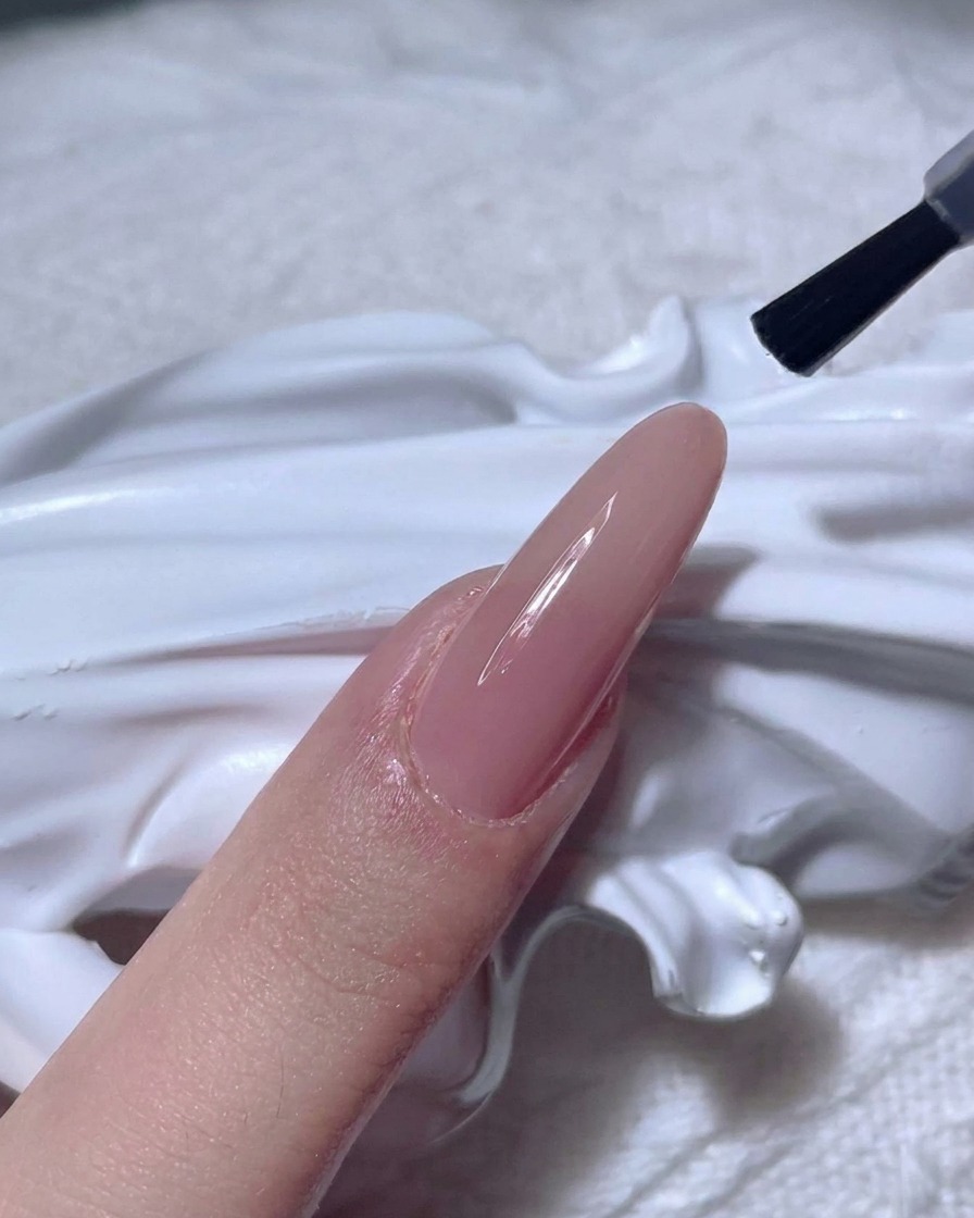 Un pincel para uñas aplicando un top coat transparente y brillante sobre una uña de color nude.