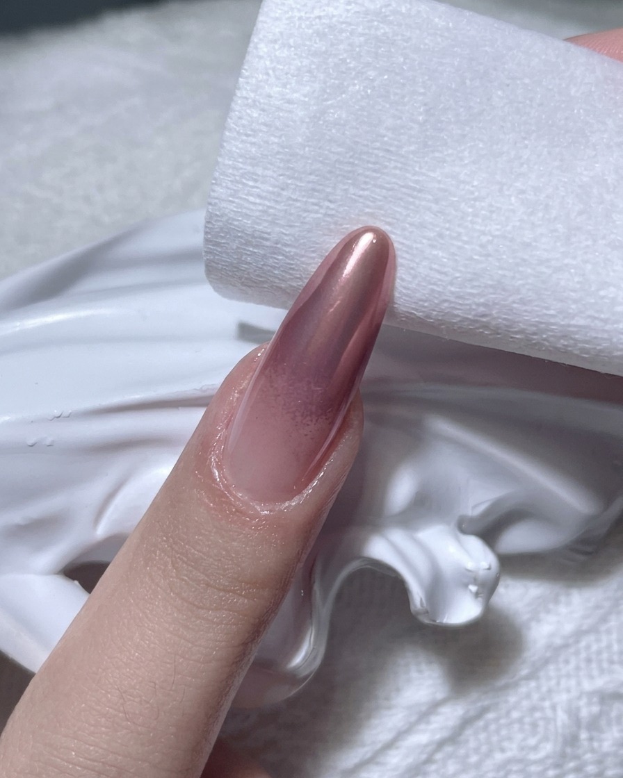 Primer plano de una mano usando una toallita sin pelusa para limpiar los bordes de una uña con acabado cromado en oro rosa.