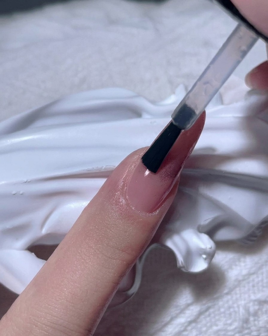 Un pincel aplicando un gel transparente y espeso sobre una uña cromada en oro rosa.