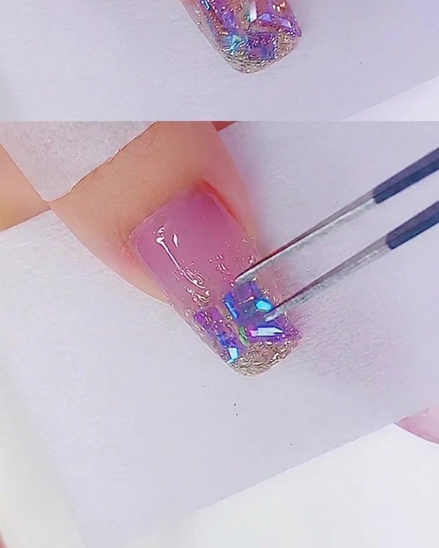 Plano detalle de unas pinzas colocando pedrería iridiscente en una punta de uña ya cubierta con gel de purpurina oro rosa.