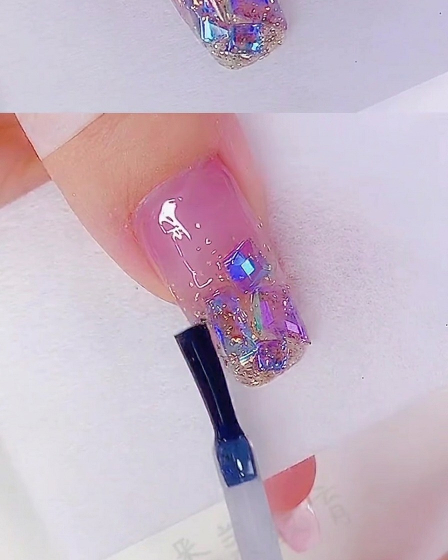 Un pincel de uñas aplicando un top coat transparente sobre una uña decorada con purpurina oro rosa y pedrería iridiscente.
