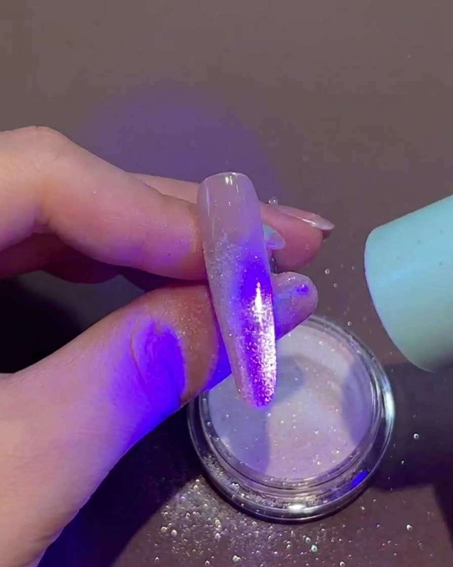 Una mano sosteniendo una pequeña lámpara UV/LED portátil directamente sobre una uña que tiene gel húmedo y polvo suelto en la punta.