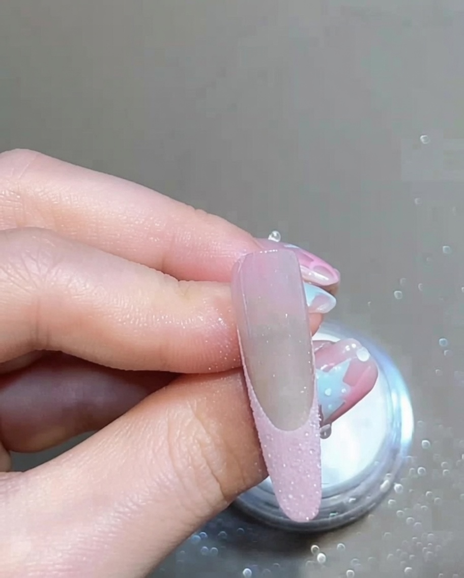 Un pincel suave cepillando una uña manicurada para eliminar el exceso de polvo brillante suelto, mostrando una punta francesa limpia.