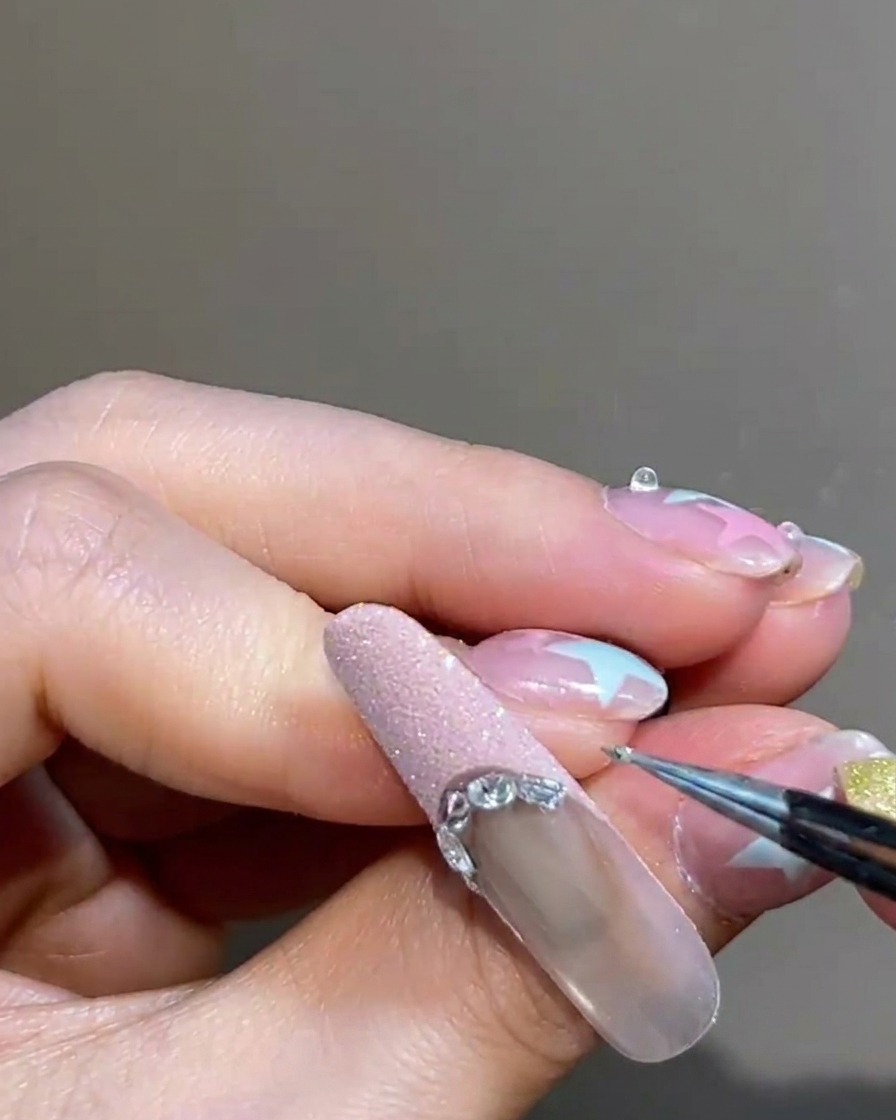 Un primer plano de unas pinzas colocando una pequeña cuenta metálica plateada en el borde de una punta francesa rosa.