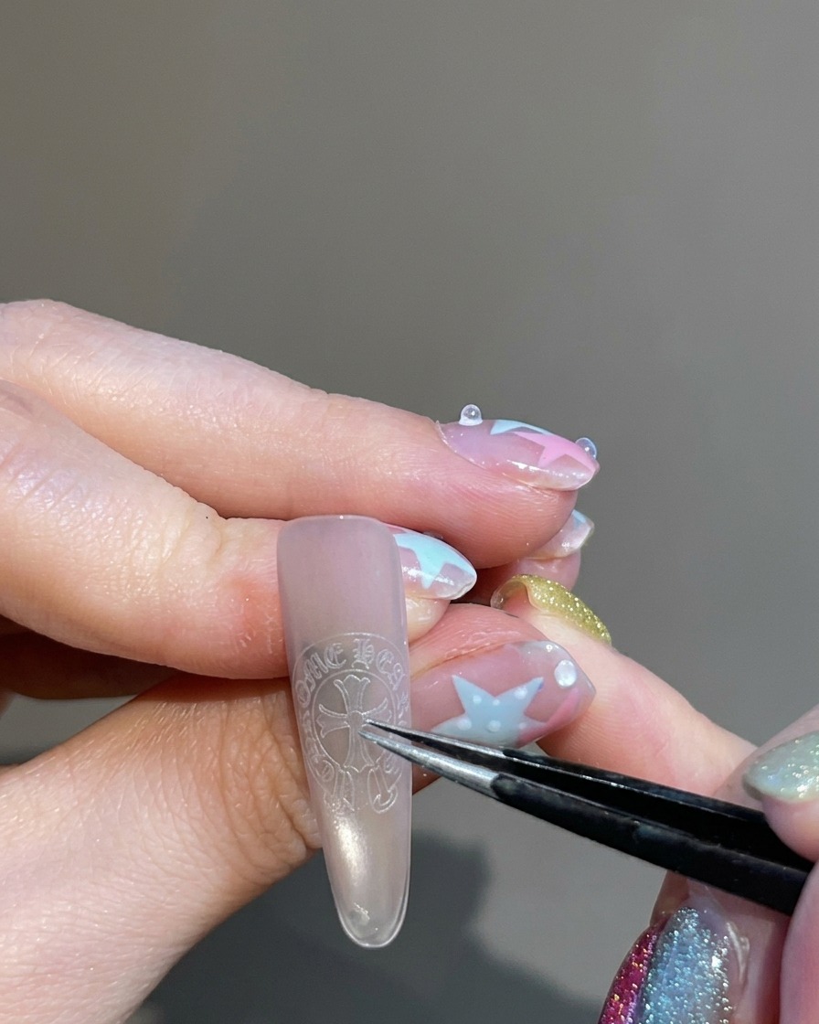 Pinzas sosteniendo y colocando una pegatina en forma de cruz en el centro de la uña.