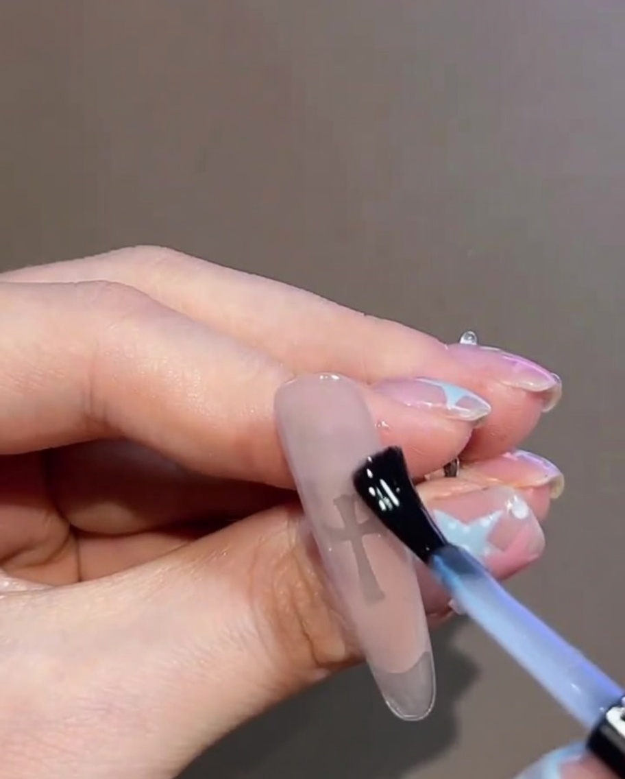 Primer plano de la aplicación de un top coat de gel transparente sobre el diseño terminado de uñas con la cruz y la pedrería.