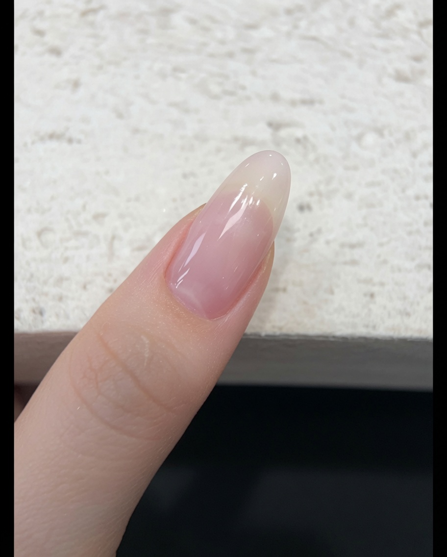 Un pincel aplicando un esmalte de gel jelly traslúcido tono nude rosado sobre una uña natural.