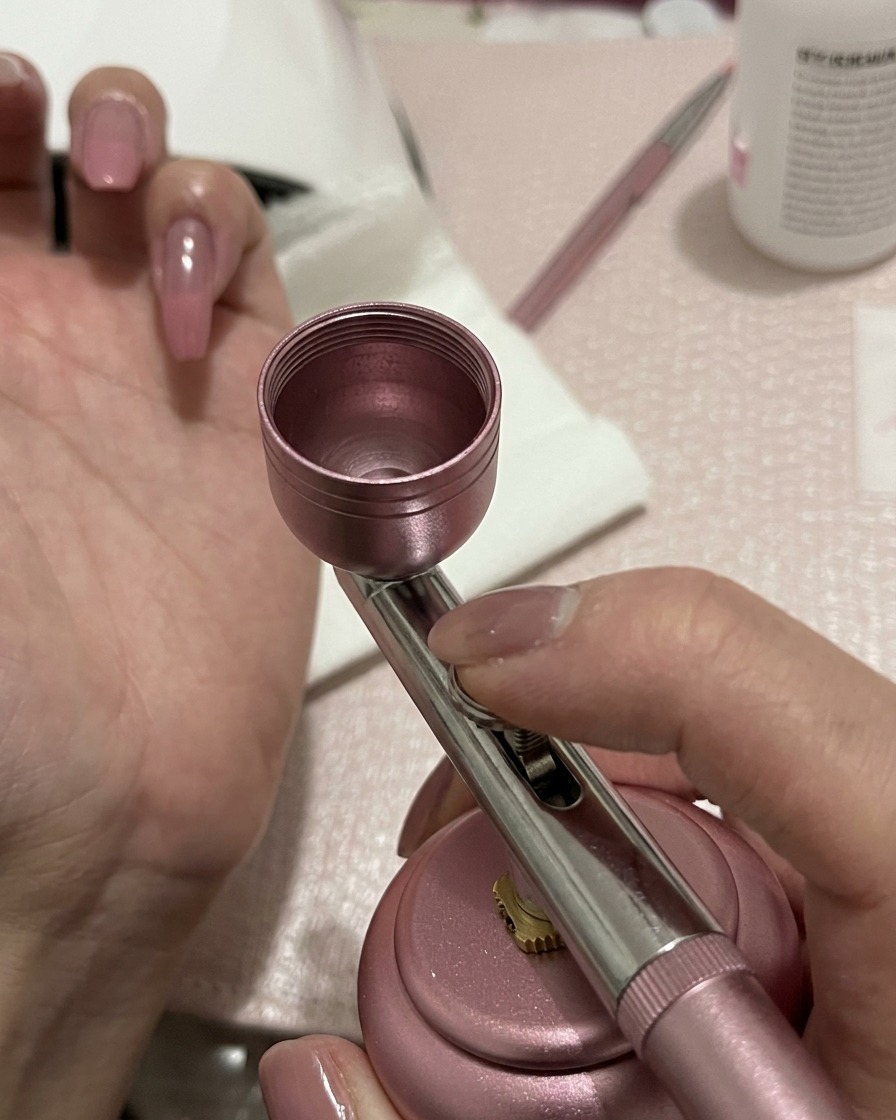Un aerógrafo aplicando una ligera capa de gel melocotón-rosa al centro de una uña preparada.