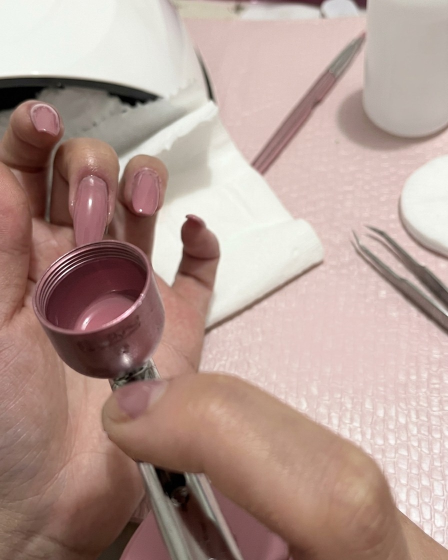 Primer plano de una mano sosteniendo un aerógrafo, rociando una fina bruma color melocotón sobre las uñas manicuradas.