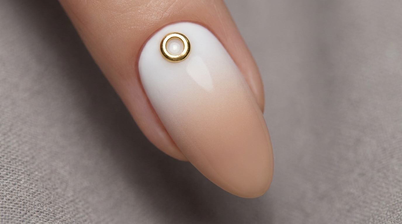 Uñas con un suave degradado aerografiado de blanco a nude, rematadas con un dije minimalista de anillo metálico dorado.