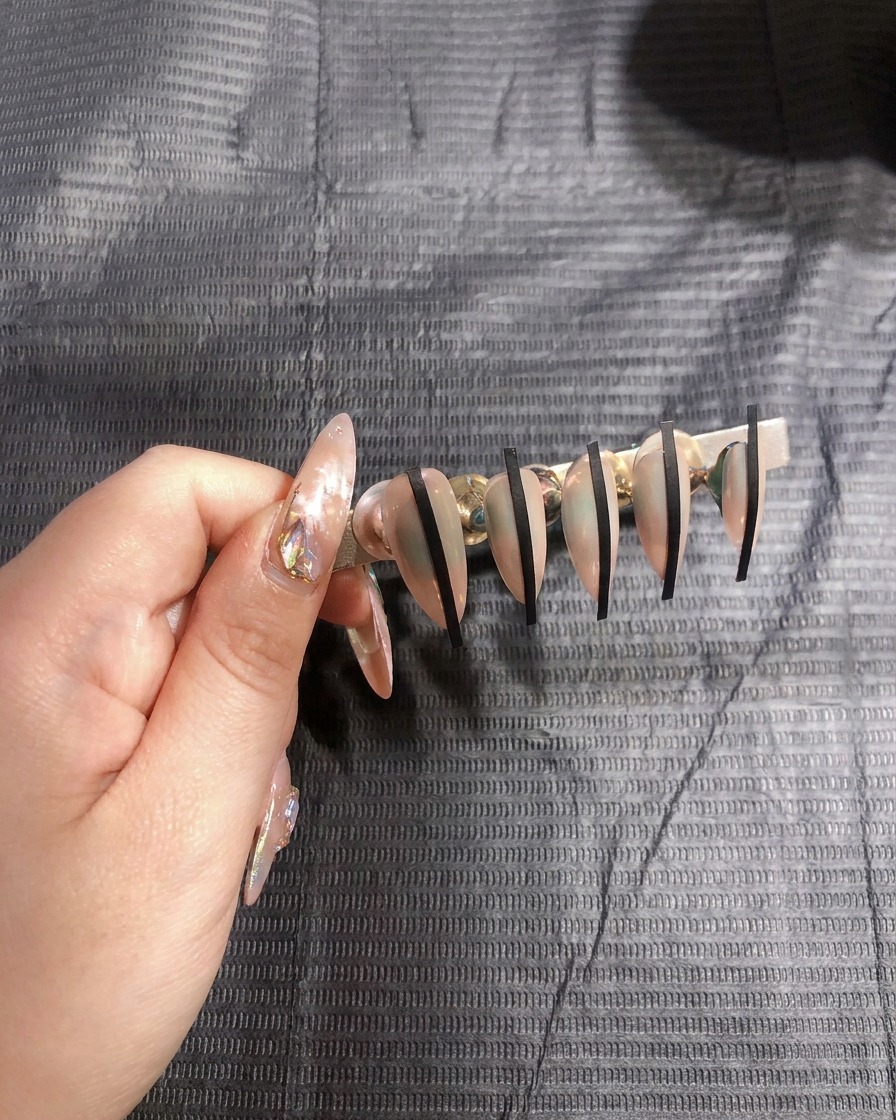 Primer plano de una mano colocando una fina pegatina negra verticalmente sobre una uña de tono nude.