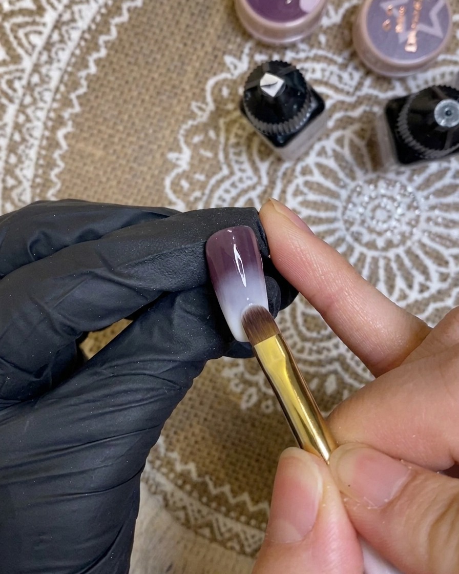 Primer plano de un pincel aplicando un gel morado oscuro y rico en la punta de una uña.