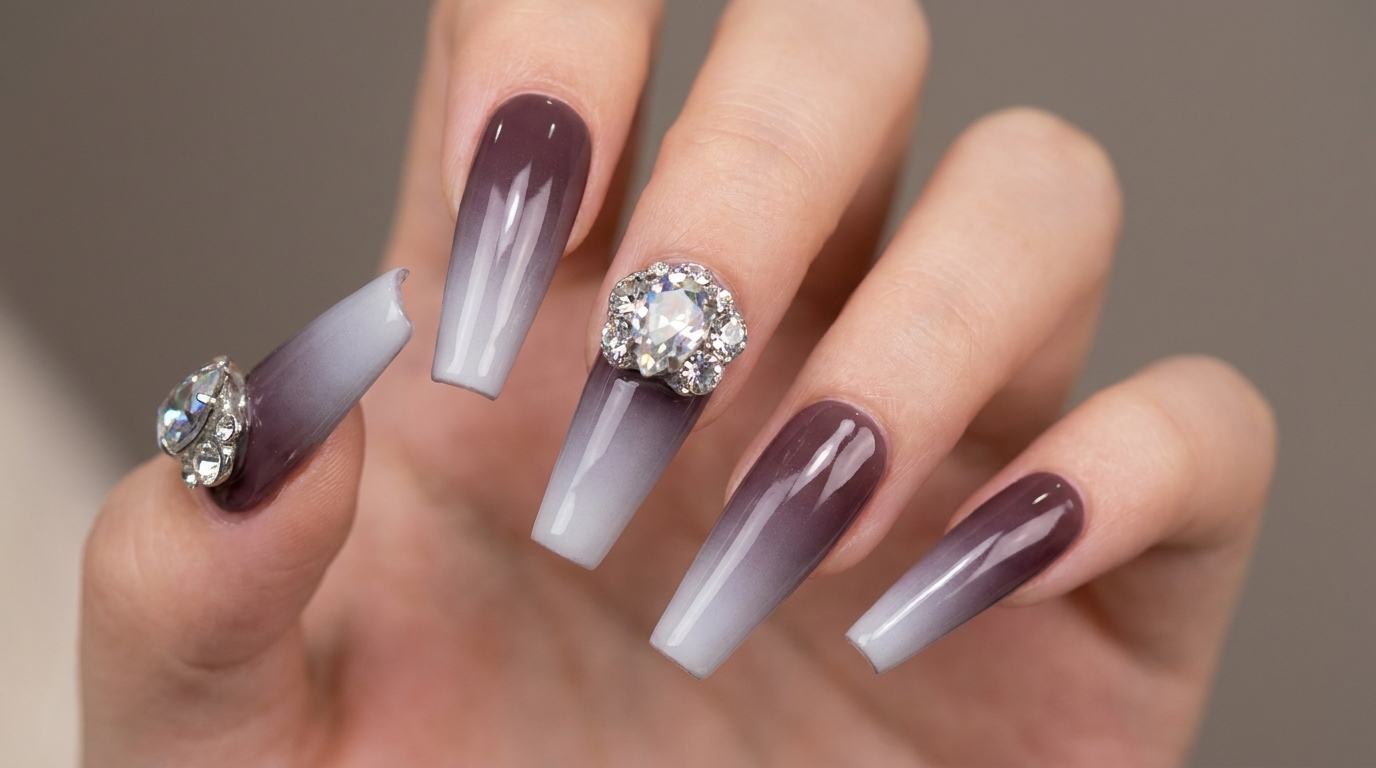 Uñas largas con forma coffin que muestran un suave degradado con aerógrafo de morado oscuro a blanco, rematadas con un elaborado diseño de pedrería.