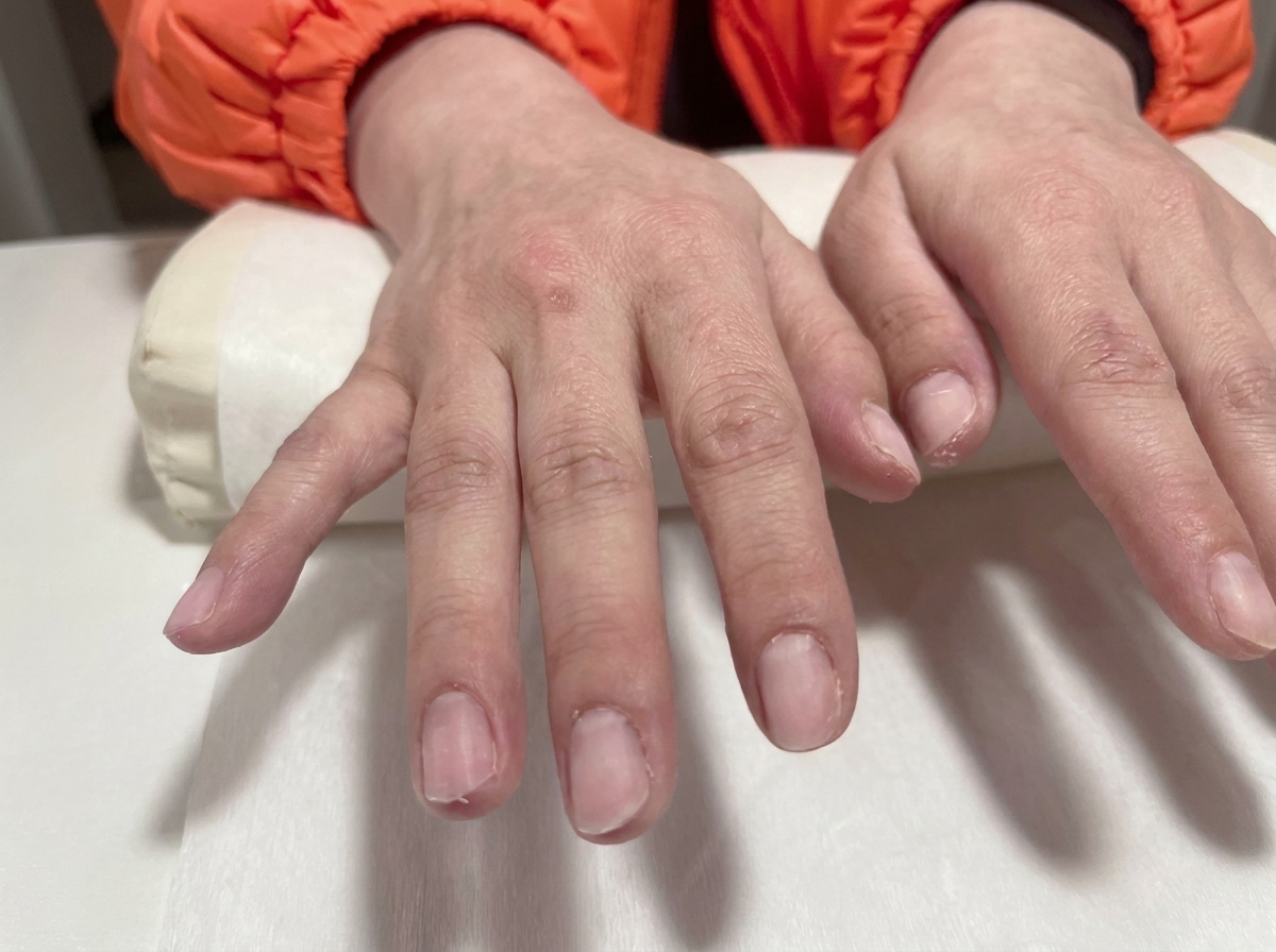 Primer plano de unas manos con uñas de gel preparadas y transparentes descansando sobre un cojín de salón blanco.