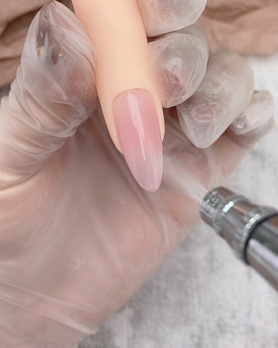 Aerógrafo aplicando un suave degradado blanco sobre la punta de una uña con base de gel transparente.