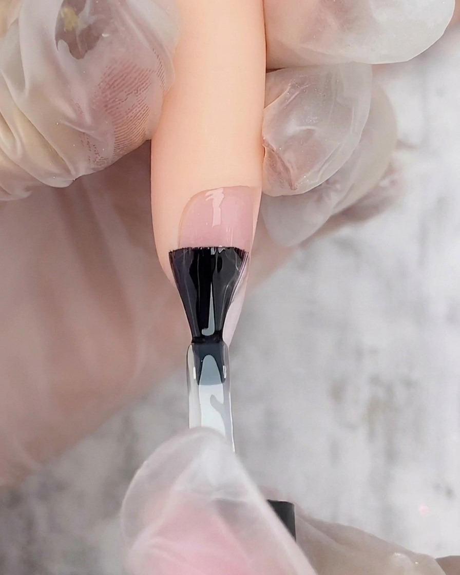 Pincel de esmalte negro aplicando una capa de top coat transparente sobre una uña ombré blanca y rosa.