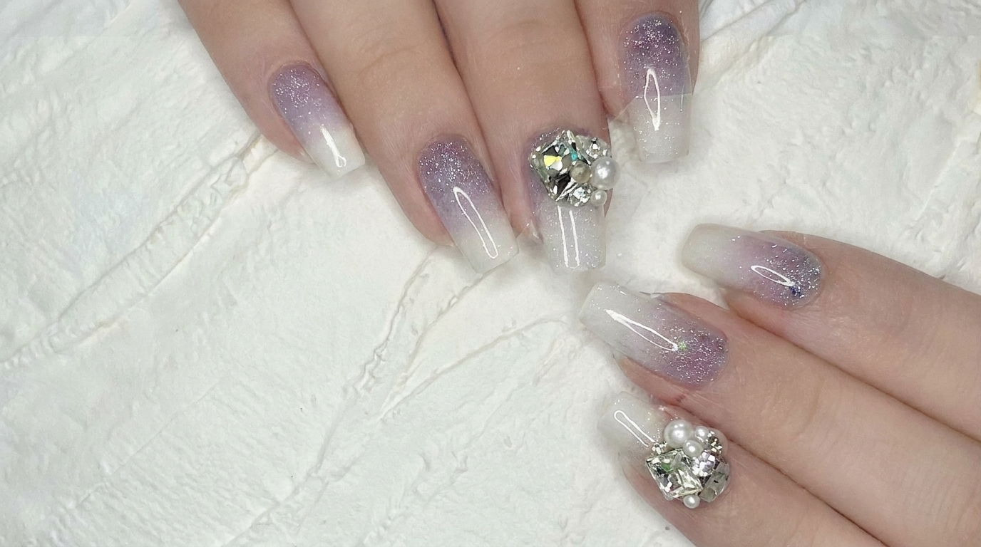 Un impresionante primer plano de uñas de gel ombré de morado a blanco hechas con aerógrafo y decoradas con delicados cristales plateados.