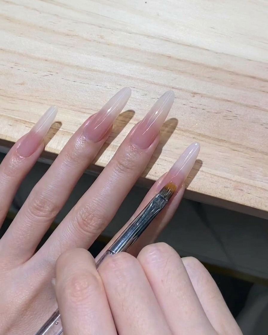 Pincel dentado para ombré arrastrando suavemente gel blanco lechoso húmedo hacia abajo en una extensión de uña stiletto transparente.