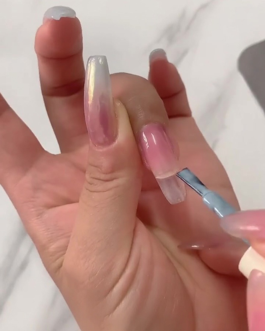 Primer plano de un pincel aplicando esmalte en gel azul claro en la mitad superior de una uña larga transparente.