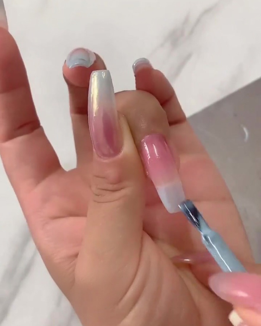 Primer plano de un pincel plano difuminando el esmalte en gel azul concentrado desde la punta hacia abajo sobre una base nude traslúcida.