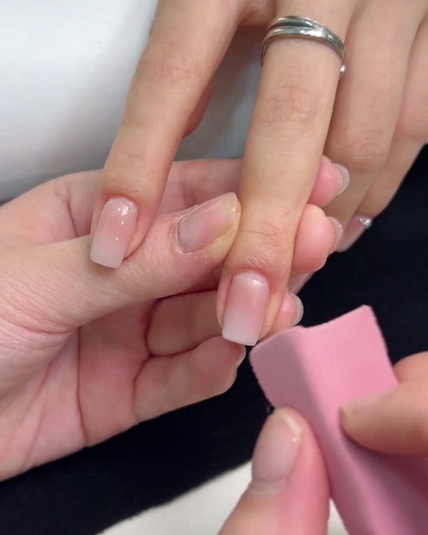 Una esponja rosa siendo utilizada para aplicar esmalte de gel blanco en la punta de una uña cuadrada, difuminándolo para crear un efecto ombré.