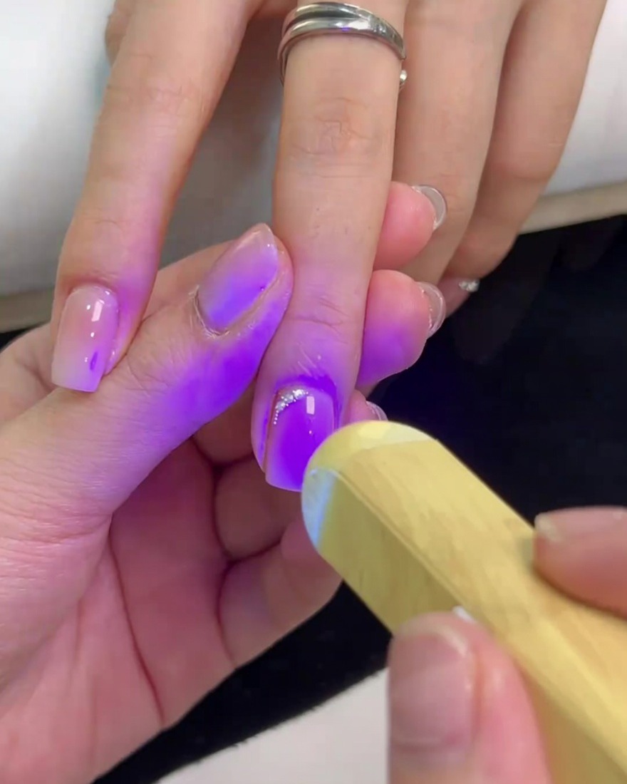 Una lámpara UV portátil brillando luz púrpura sobre una uña cuadrada para curar el pegamento y asegurar los cristales.