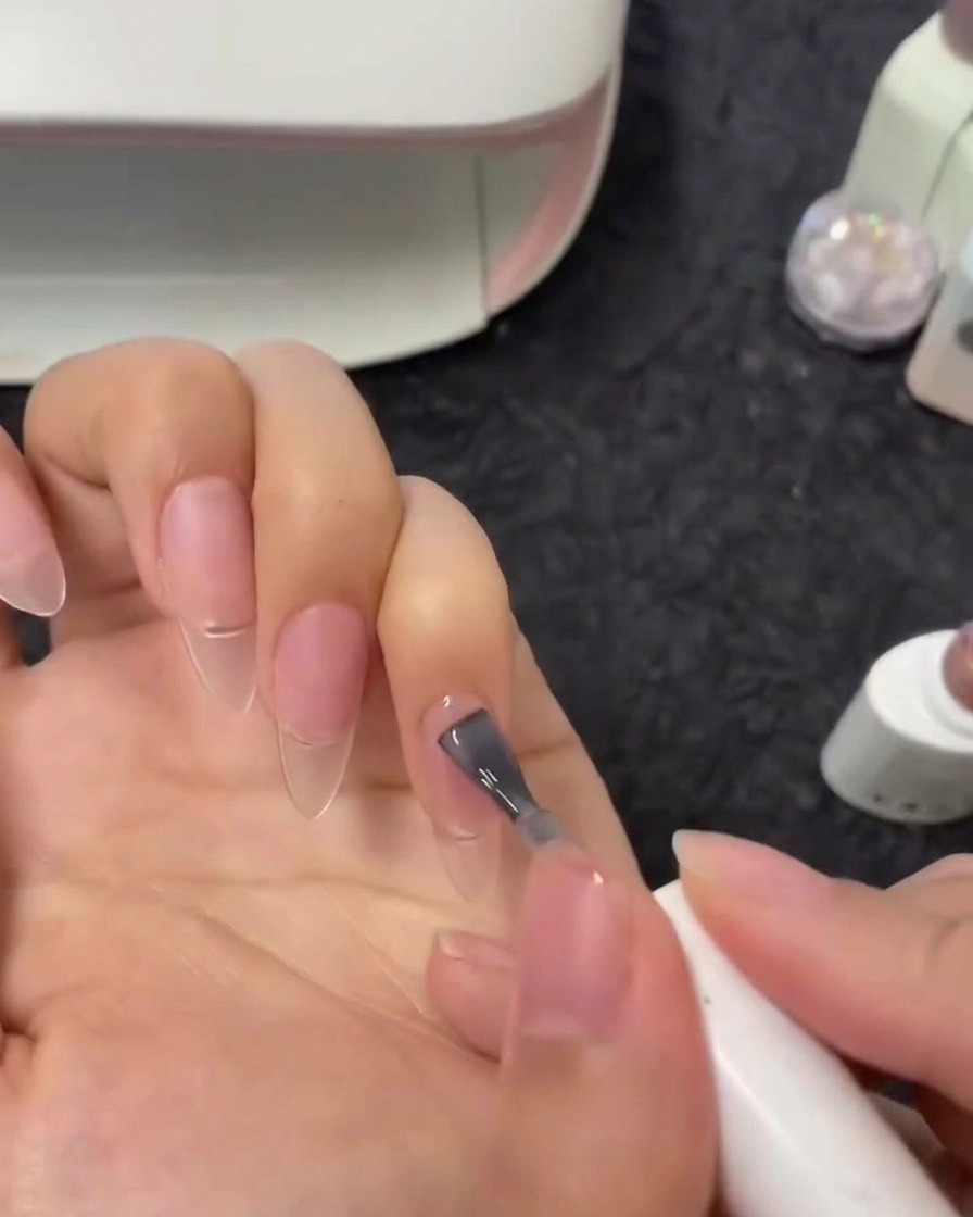 Vista en primer plano de un pincel aplicando esmalte de gel nude semitransparente sobre una uña índice.